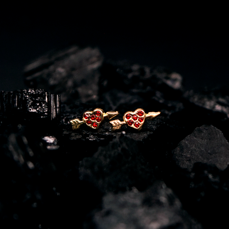 Broquel con forma de Corazón flechado con Zirconias rojas en Corte Brillante.  Oro Amarillo de 10 kilates.